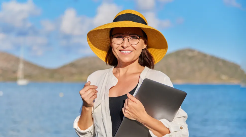 photo of woman standing in front of scenic water and mountain view wearing a sunhat and holding a laptop