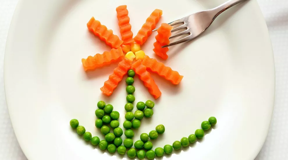 Peas, carrots, and corn arranged on a plate to look like a flower. A fork reaches for one of the carrots. 