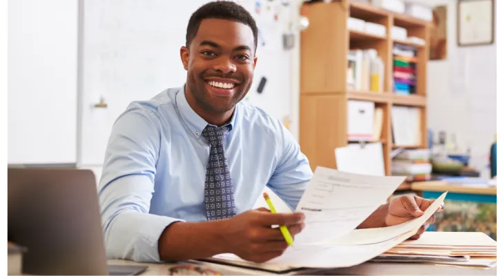 photo of male teacher smiling at desk with laptop and papers