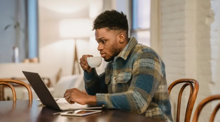 photo of a man sitting at a table wearing a warm jacket looking at a laptop and drinking something warm