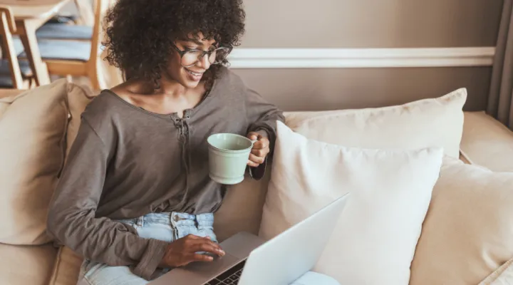 photo of woman sitting on a couch holding a coffee while looking at a laptop